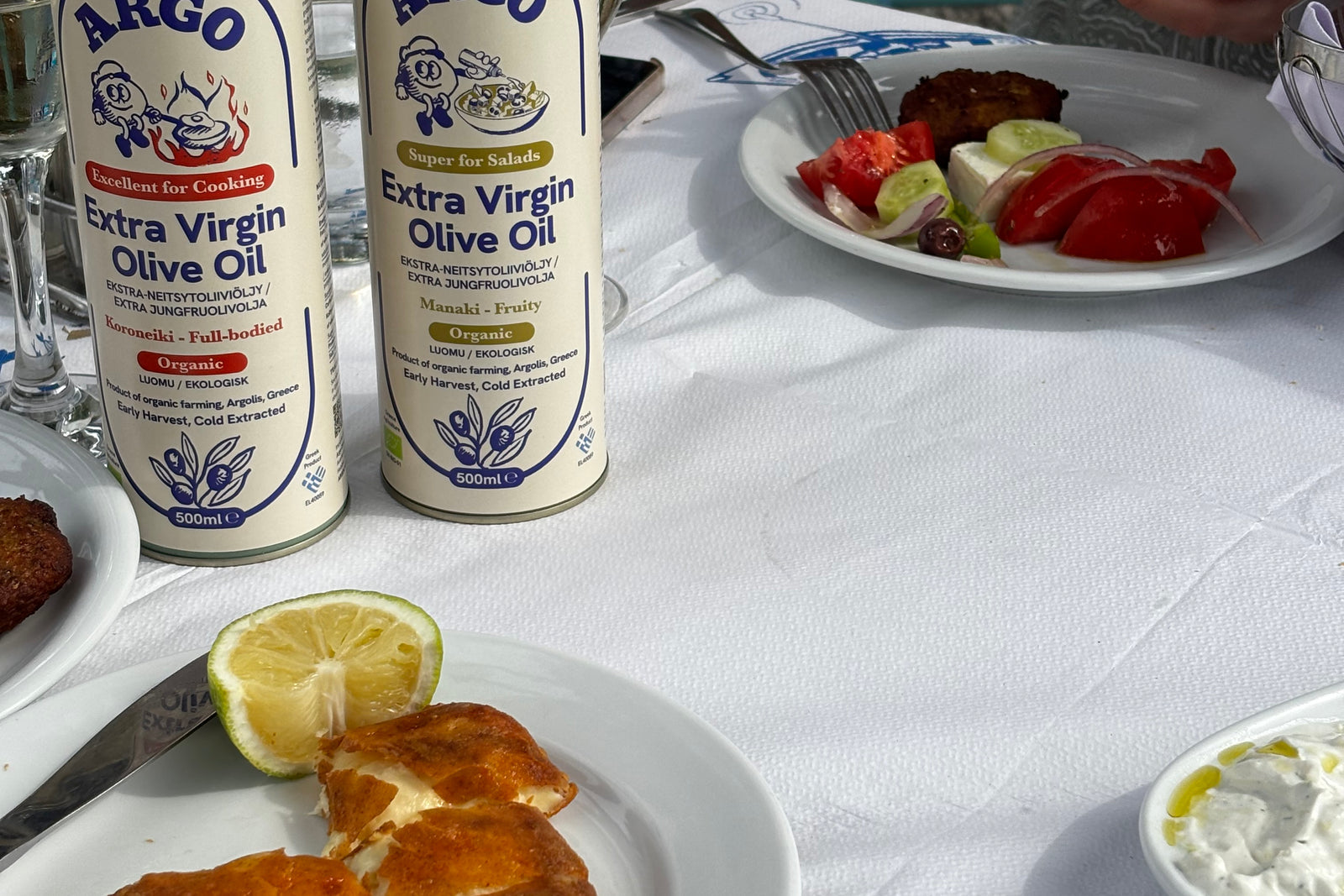 Two tins of Argo olive oil on a taverna table beside fried saganaki and lemon.
