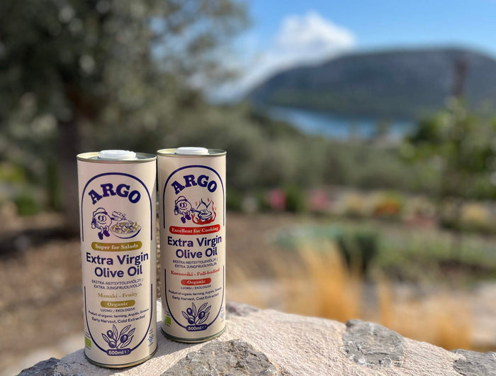 Two cans of Argo Extra Virgin Olive Oil on a stone surface with a scenic background.